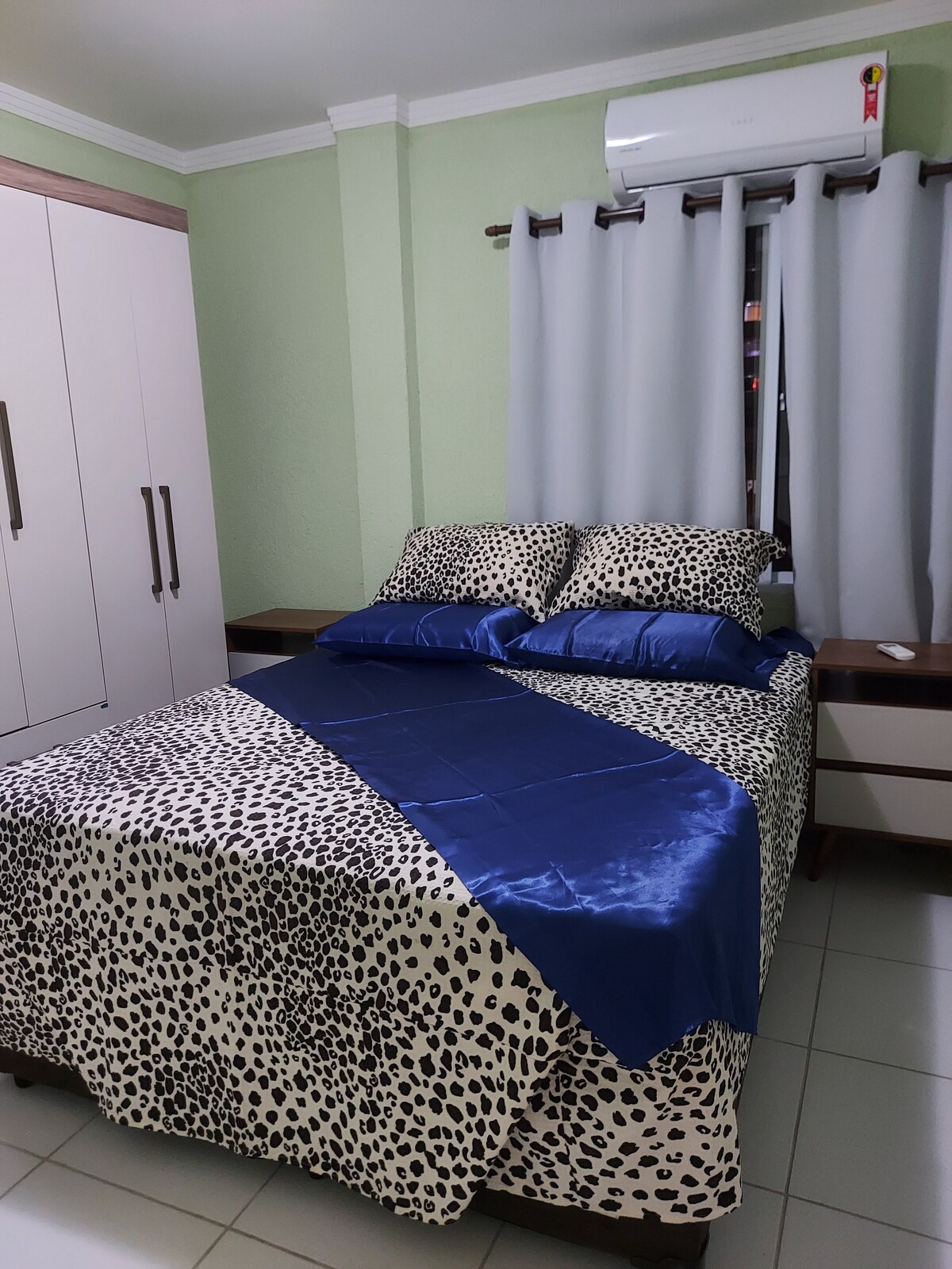 A cozy sleeping area features a spacious bed with a patterned comforter and coordinating decorative pillows. Light grey curtains frame the window, allowing natural light to fill the space. The room is equipped with a wardrobe and bedside tables, complemented by a calming green wall.