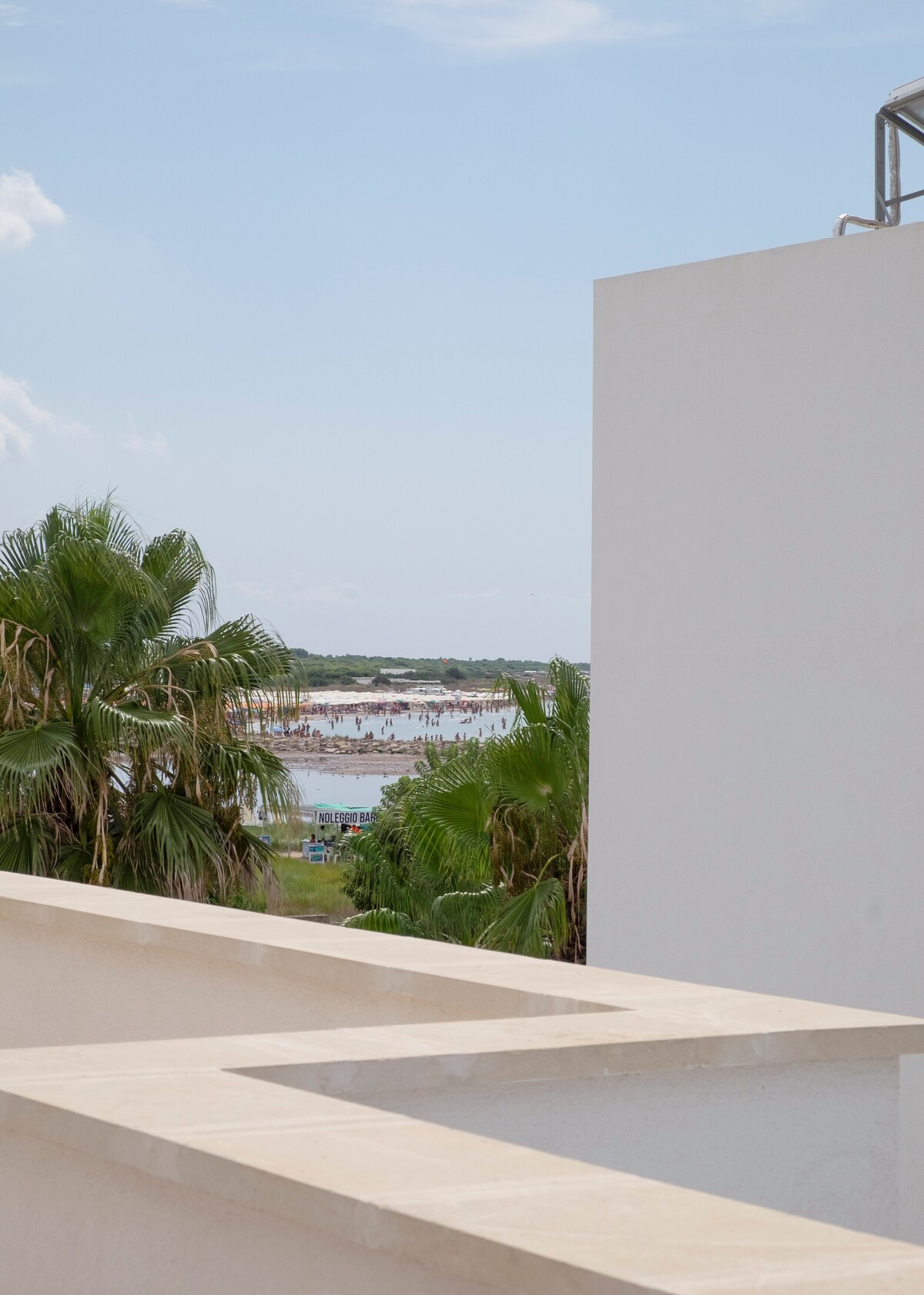 A serene view of the nearby beach and vibrant palm trees is captured from a balcony. The white structure of the building complements the natural surroundings, while the calm water reflects the sunny sky in the distance.