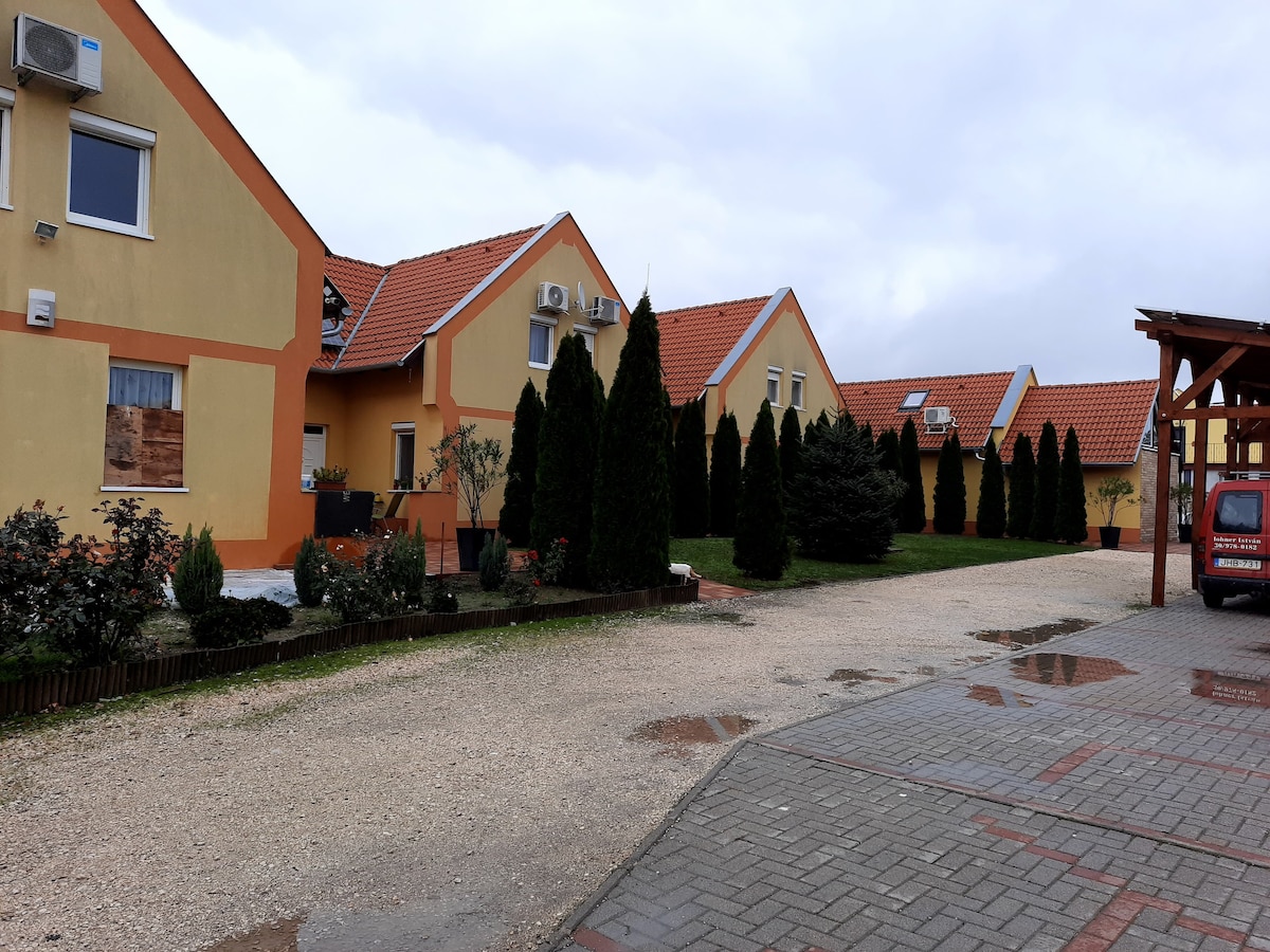 A row of yellow buildings with red roofs is visible, accompanied by neatly trimmed evergreen shrubs. A gravel pathway leads to the entrance of the buildings, with a parking area seen to the right. The sky is overcast, contributing to a calm ambiance.
