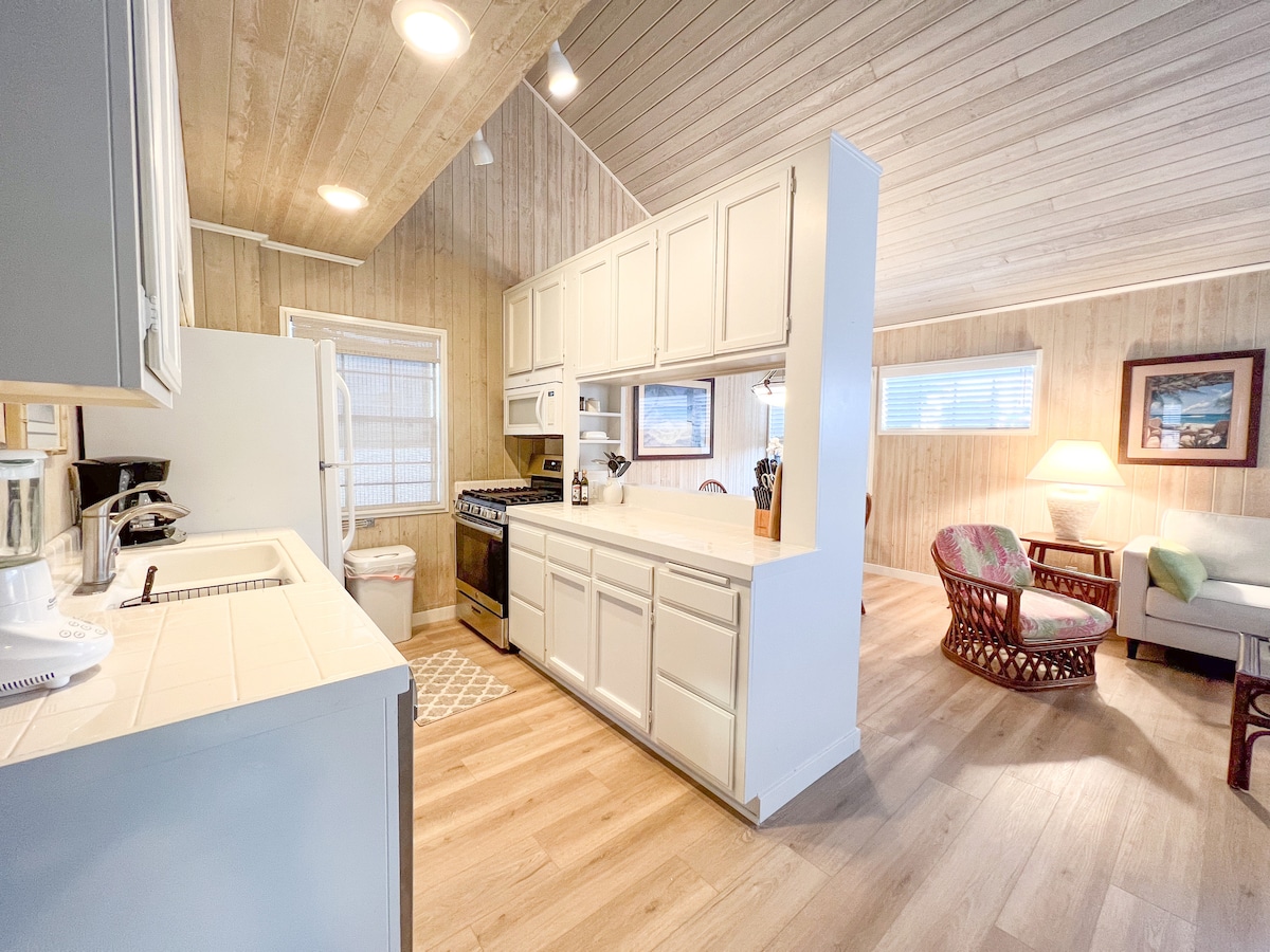 A spacious kitchen features white cabinetry and a center island, with a gas stove and a refrigerator visible. Natural light fills the room, illuminating the cozy seating area nearby, which includes a wicker chair and a light-colored sofa. Wooden ceilings add warmth to the space.