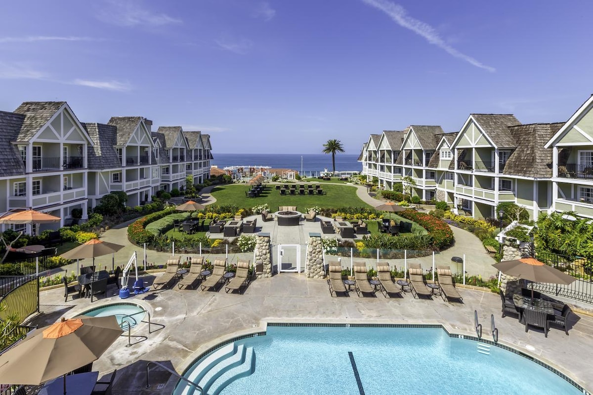 The expansive outdoor area features a swimming pool surrounded by lounge chairs. Lush gardens and neatly arranged pathways lead to nearby units, showcasing a vibrant landscape with colorful flowers. The Pacific Ocean is visible in the distance, enhancing the coastal ambiance.