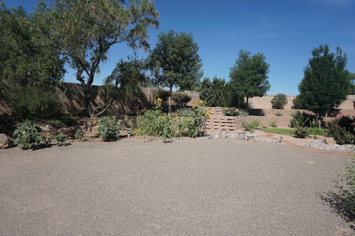 An outdoor space is presented, featuring a landscaped area with a variety of plants, including sunflowers. The surrounding trees provide shade, while a stone staircase leads to a higher elevation in the background. The ground is composed of gravel, enhancing the natural feel.