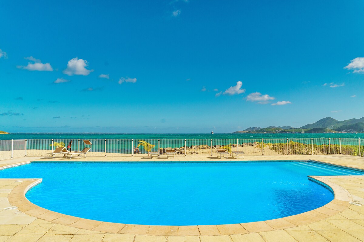 A large circular swimming pool with clear blue water is set against a backdrop of the ocean. Sun loungers are positioned along the edge of the pool area, providing a space for relaxation with views of the shoreline and hills in the distance. The sky is bright and blue.
