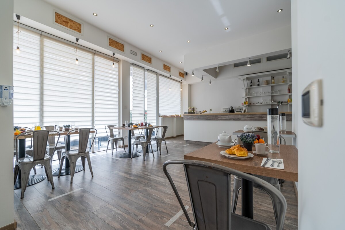 A bright and airy dining area is equipped with modern furnishings. Several tables are set for guests, with breakfast items displayed, including fruit and pastries. Large windows allow natural light to fill the room, enhancing the welcoming atmosphere.