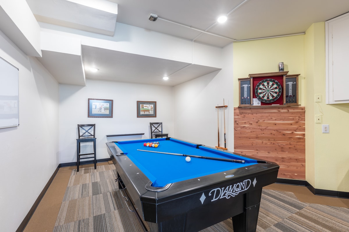 A game room is presented featuring a pool table with colored balls arranged on it. Opposite the table, a wall-mounted dartboard is displayed, surrounded by framed artwork. Two chairs are positioned nearby, and the space is finished with a beige and grey striped carpet.