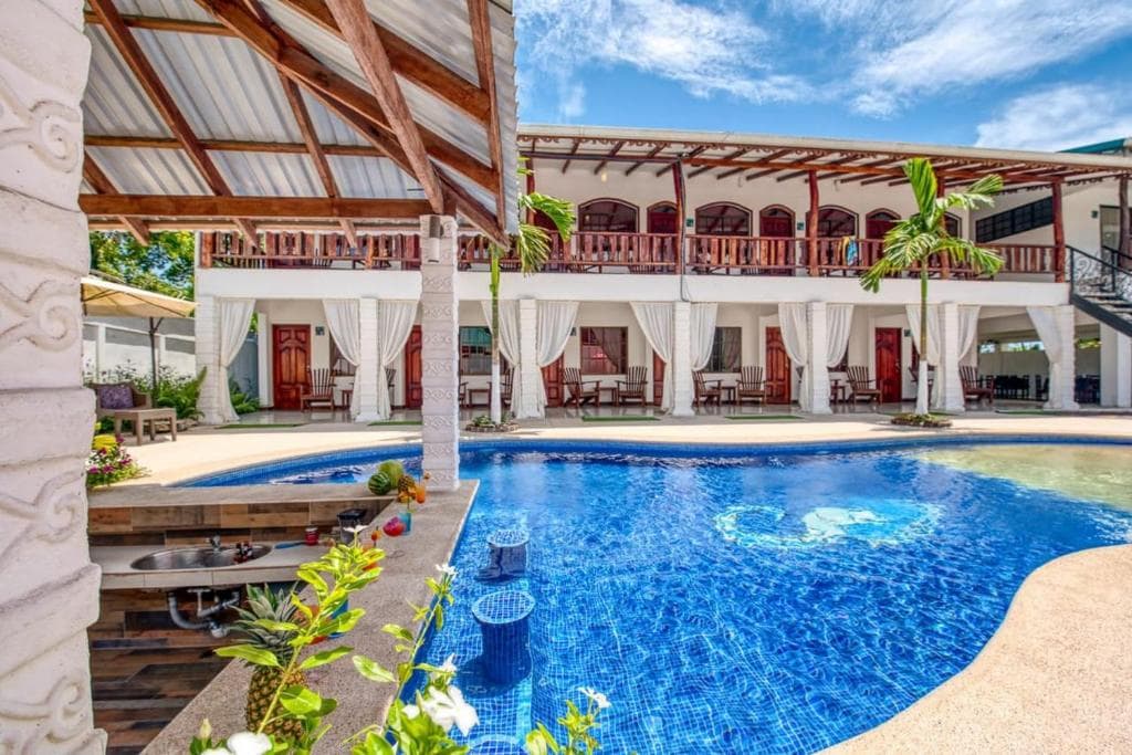 An outdoor area features a large pool surrounded by lounge chairs and greenery. The building's upper floor displays multiple balcony spaces with wooden railings, while curtains softly drape the ground-level areas, creating a blend of relaxation and elegance.