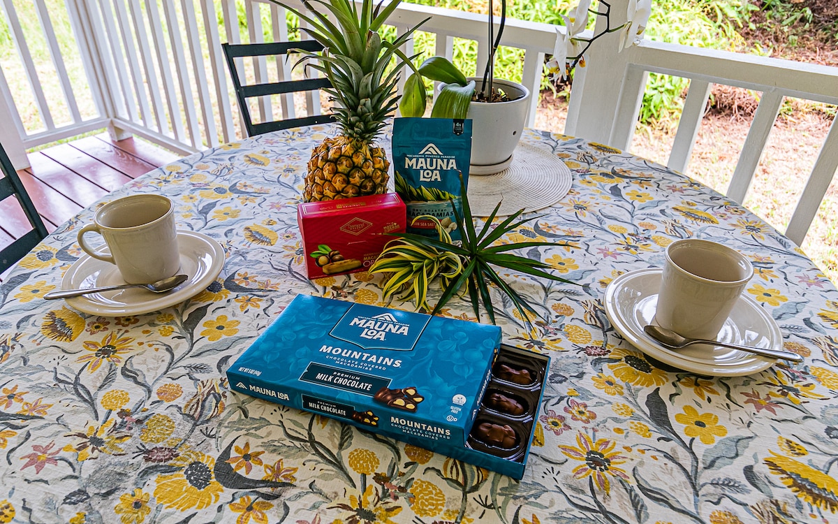 A round table is set with two beige cups and a white tablecloth adorned with floral patterns. A box of chocolates and a bag of snacks are positioned at the center, alongside a pineapple and decorative foliage, highlighting a pleasant gathering spot.