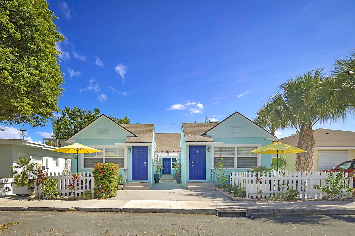 Two charming bungalows feature pastel blue exteriors, white picket fencing, and yellow umbrellas providing shade. Lush greenery surrounds the pathway leading to the entrances, enhancing the appealing outdoor space. A clear blue sky adds vibrancy to the scene, complementing the coastal aesthetic.