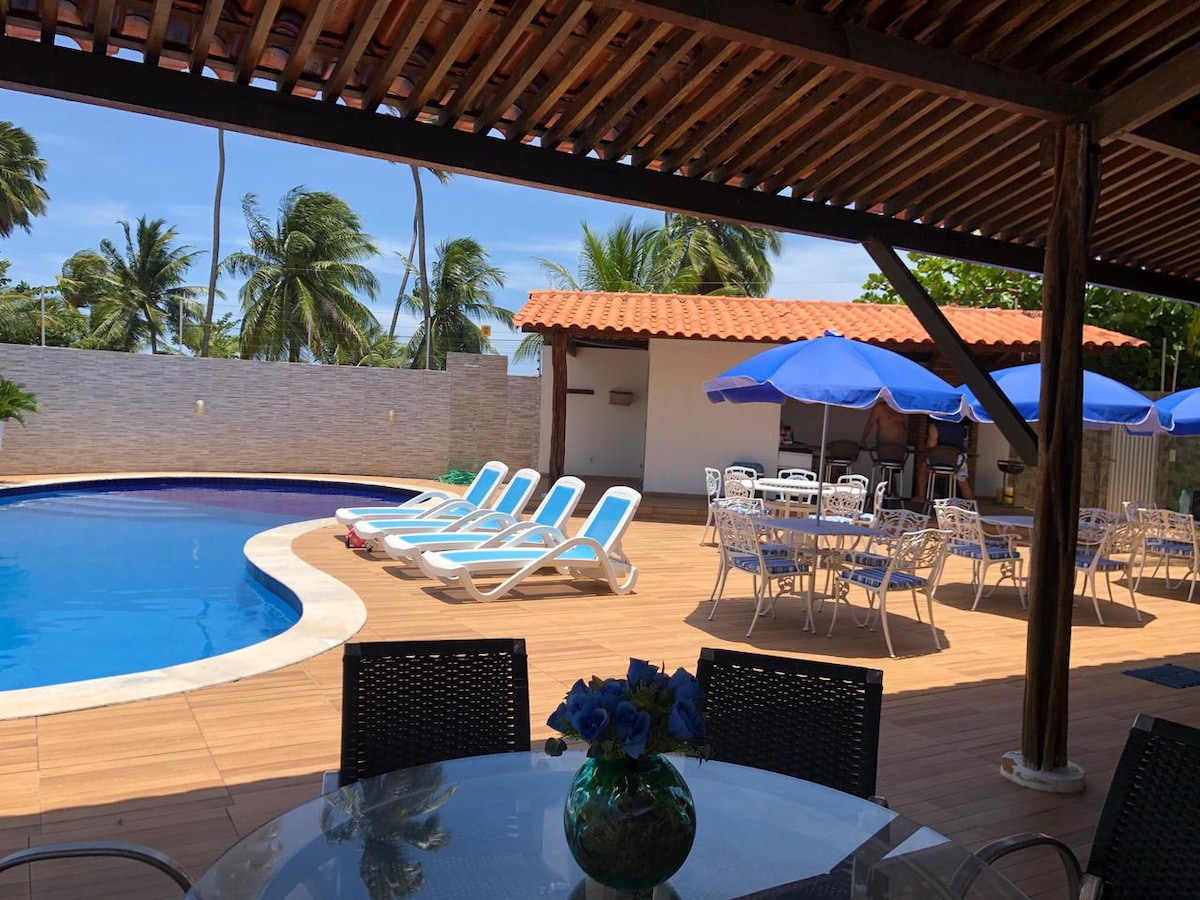 A spacious outdoor area features a swimming pool surrounded by lounge chairs. A covered dining space with tables and chairs is positioned nearby, complemented by shaded umbrellas. Lush palm trees in the background provide a tropical setting.