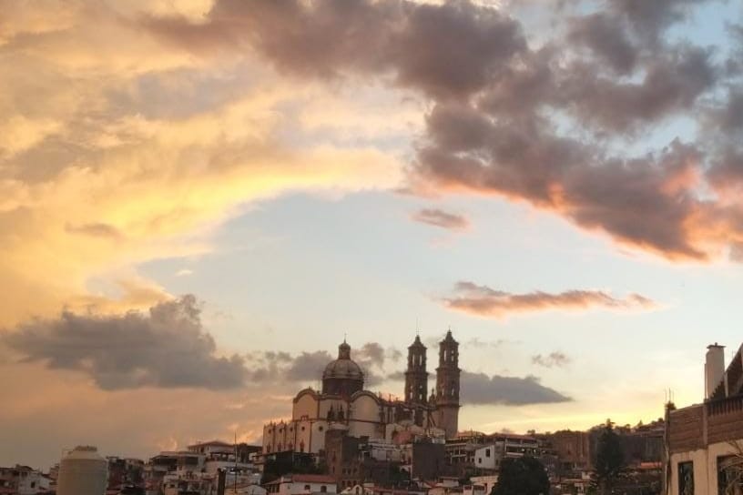 A panoramic view of a tranquil sunset casts warm hues over the skyline of Taxco, with its distinctive architecture highlighted against the colorful clouds. The silhouette of church domes and bell towers can be seen, providing a glimpse of the area's historical charm.