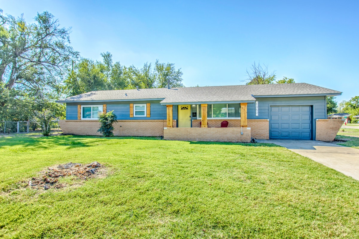 A single-story residence is positioned on a spacious lawn, featuring a well-maintained exterior with a blend of blue siding and brick accents. A covered porch welcomes visitors, while a driveway leads to an attached garage on the right side.