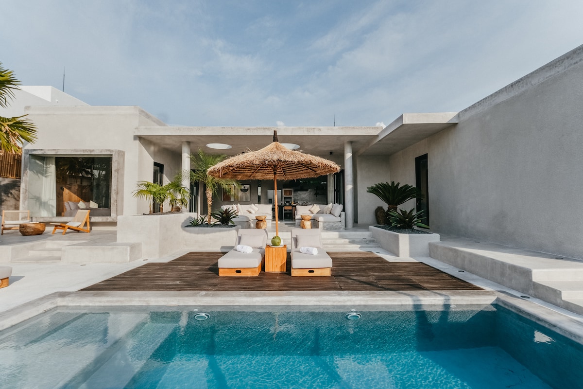 An outdoor area showcases a private pool surrounded by a wooden deck and minimalistic sun loungers. A thatched umbrella provides shade, while tropical plants enhance the space. The villa's modern design, featuring a mixture of concrete and wood, creates a serene environment for relaxation.