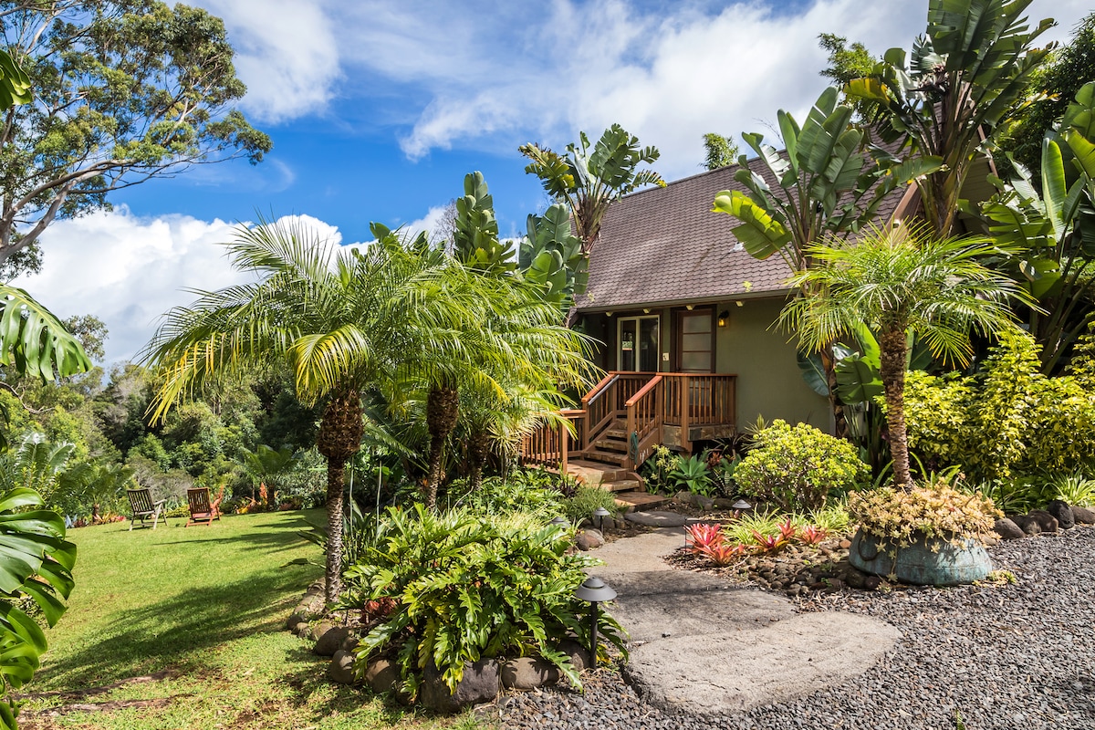 A charming cottage is nestled among tropical greenery, featuring lush palms and vibrant plants. The entrance is highlighted by a welcoming staircase leading up to a covered porch. A scenic landscape can be observed in the background, showcasing the natural beauty of the surroundings.