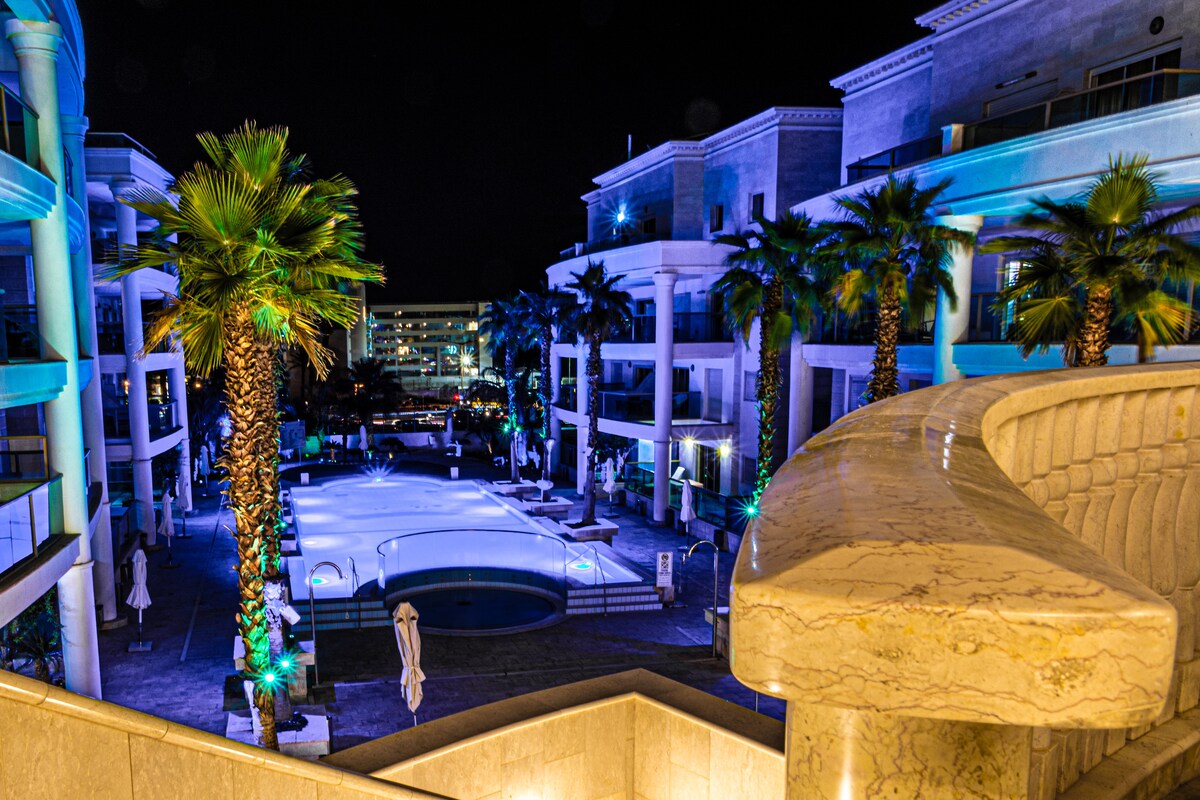 An evening view of a private luxury complex is captured, featuring a well-lit pool area surrounded by palm trees. Soft blue and green lighting enhances the modern architecture, while lounge chairs and umbrellas are arranged neatly around the pool.