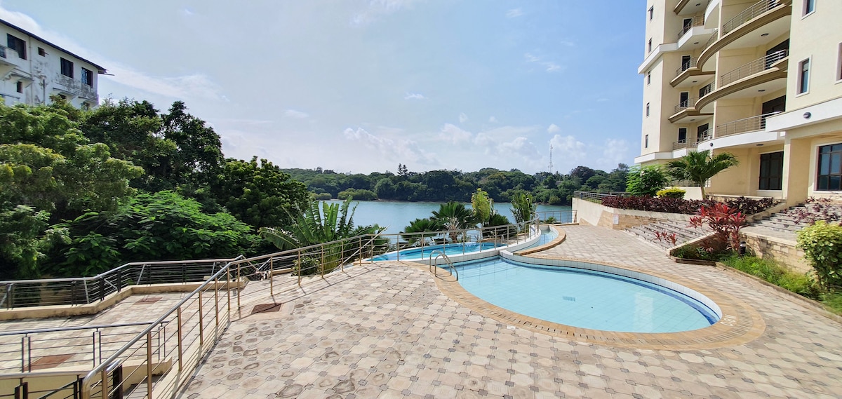 A swimming pool is framed by a spacious tiled area, with lush greenery visible beyond. The calm water reflects the clear sky, while a walking path leads along the poolside toward a tranquil body of water.