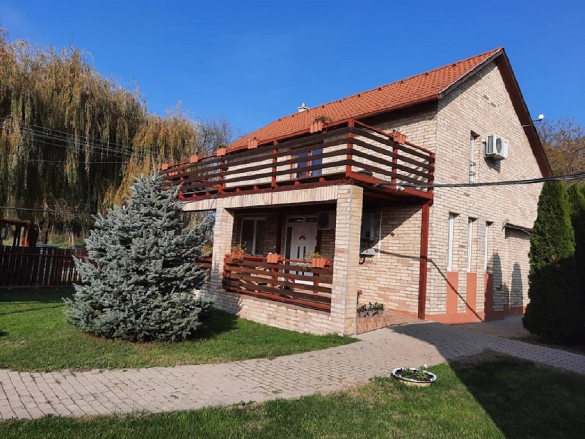 A two-story brick house is framed by a green lawn and plants. A wooden balcony with a railing extends from the upper floor, complementing the exterior. The clear blue sky enhances the overall appearance, while a paved pathway leads to the entrance.
