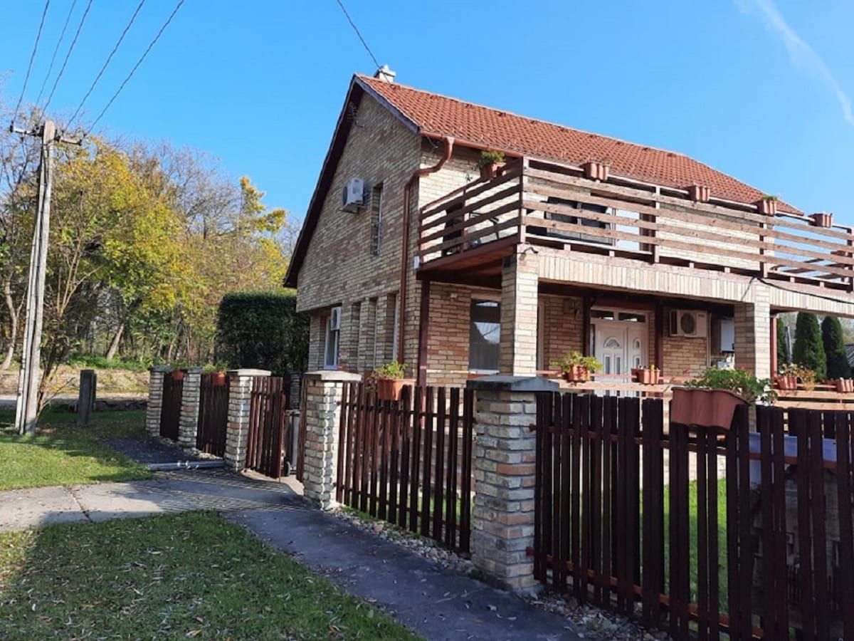 The exterior of a two-story brick house is shown, featuring a red-tiled roof and a welcoming front porch. A wooden fence surrounds the property, with potted plants along the railing. Lush greenery and trees are visible in the background, indicating a serene environment.