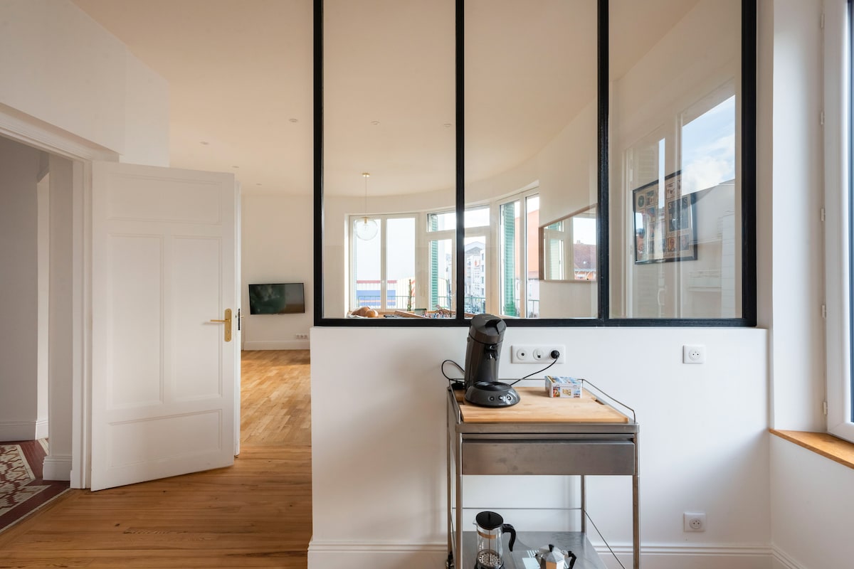 A modern kitchenette is visible with a sleek coffee machine positioned on a wooden cart. The space is illuminated by natural light streaming through large windows, showcasing a view into a connected living area featuring a television and a gentle curve of the walls.