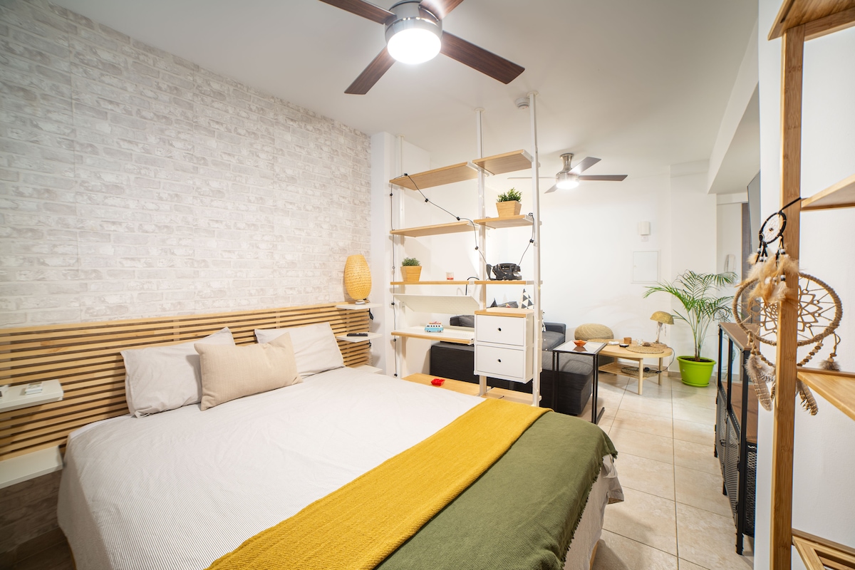 A modern bedroom features a spacious bed with neutral-colored linens and a decorative yellow blanket. A textured white brick wall complements open shelving adorned with plants and decor. Soft lighting is provided by a ceiling fan and an additional light fixture.