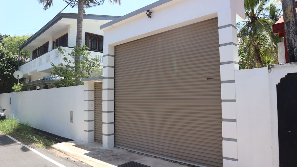 The exterior view highlights a modern holiday home featuring a two-story structure with a clean, white facade. A large, rolled-up garage door complements the sleek design. Palm trees and greenery are visible, adding a touch of nature to the inviting curb appeal.