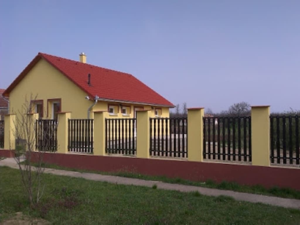 A yellow house with a red roof is set behind a wooden fence. The surrounding garden area features a well-maintained lawn, while a pathway leads to the entrance. The sky above is clear, reflecting a calm and open atmosphere.
