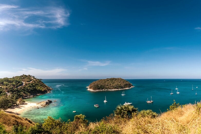 A panoramic view of a serene coastline is presented, showcasing clear turquoise waters and lush green hills. Small boats can be seen anchored in the water near a gentle island, under a bright blue sky scattered with a few clouds.