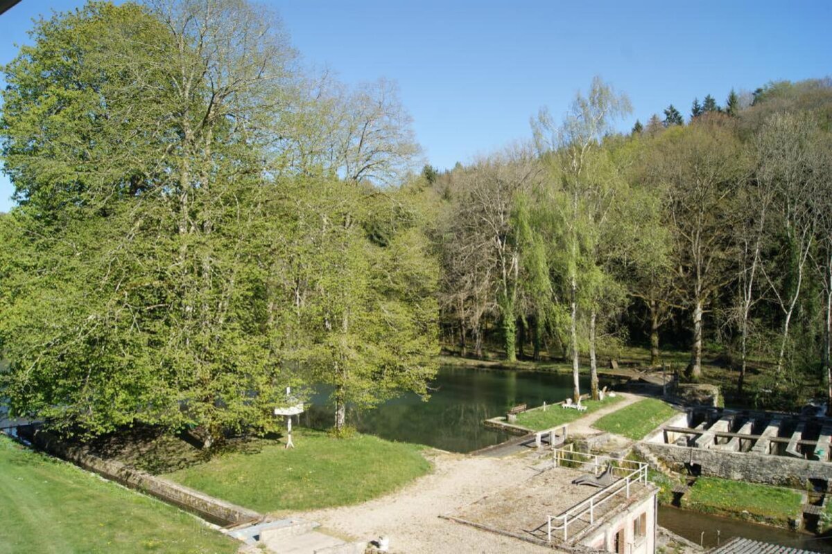 A serene landscape featuring a tranquil water body surrounded by lush greenery. Tall trees provide shade, while stone structures line the water's edge, reflecting a peaceful rural setting. The area is accessible, inviting walks along the bank.