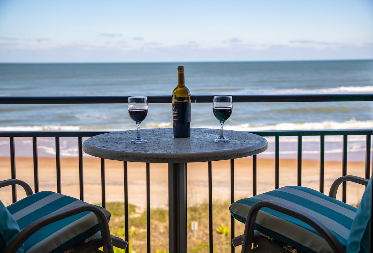 A small round table is set on the balcony, featuring two glasses of red wine beside a bottle. The ocean stretches out in the background, with gentle waves visible against the sandy shore.