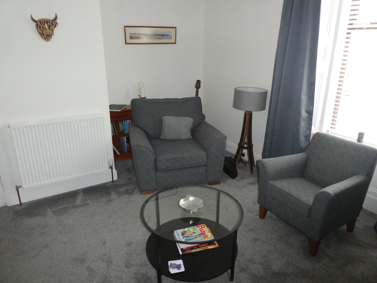 A comfortable lounge area features two upholstered chairs and a glass coffee table, positioned on a plush carpet. A small lamp and a guitar are placed nearby, while a console with books is visible against the wall. A framed image hangs above the seating area.