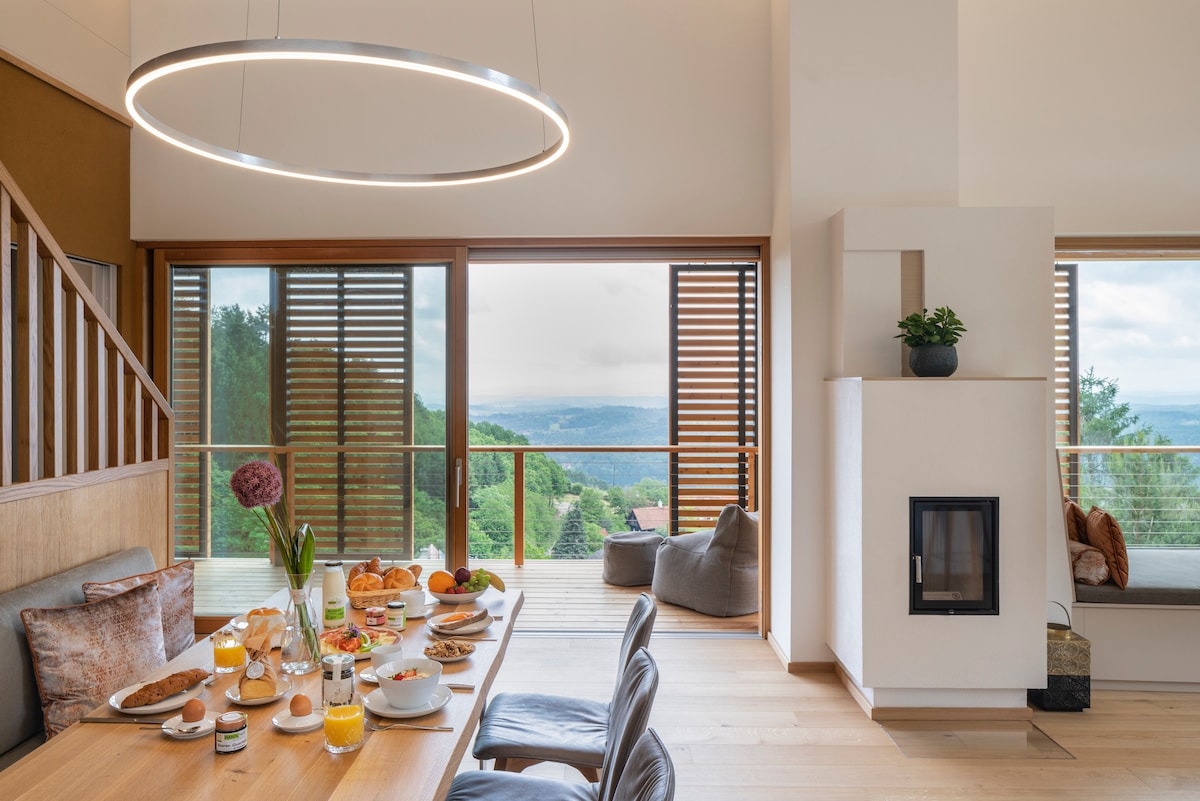 An airy dining area features a wooden table set with fresh breakfast items, complemented by orange juice and coffee. Large windows provide a panoramic view of the landscape, while a modern light fixture hangs above, and a cozy seating area with a fireplace is visible in the corner.