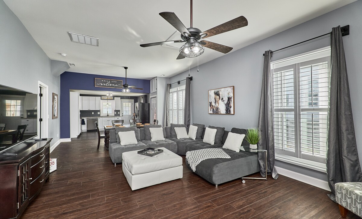 A spacious living room features a large sectional sofa in soft gray tones. Natural light enters through plantation shutters on the windows, illuminating the dark wood flooring. A modern ceiling fan hangs above, and a television is positioned on a dark wooden cabinet in the corner.