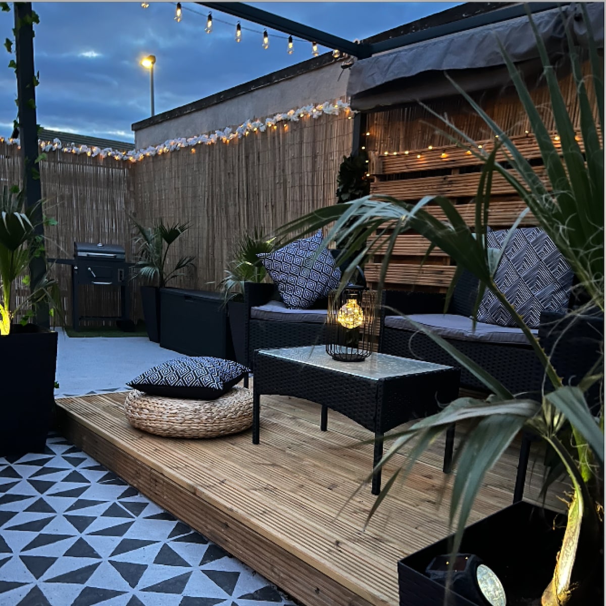 A cozy outdoor seating area is featured on a wooden deck, surrounded by potted plants. Soft lighting creates an inviting ambiance, with decorative string lights overhead. Comfortable seating is arranged, and a small table is positioned in the center, perfect for alfresco dining.