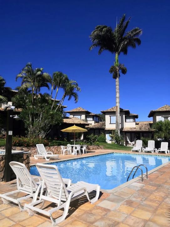 A clear blue sky frames a tranquil swimming pool surrounded by loungers under the shade of palm trees. The pool area features vibrant greenery and a sun umbrella, with charming multi-story houses visible in the background.