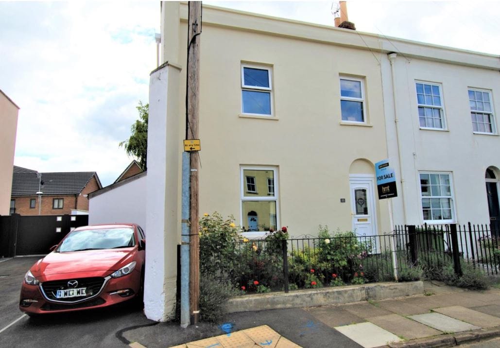 A corner property is depicted, featuring a cream-colored facade with a welcoming front door. A red car is parked in the designated area, while flower beds with plants are visible along the low fence.