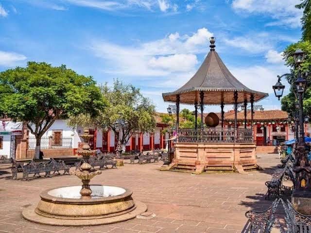 A charming public square is featured, with a decorative gazebo at its center surrounded by lush green trees. Benches are positioned around the square, and an ornate fountain adds an inviting element. Traditional architecture is visible in the background, enhancing the local character.
