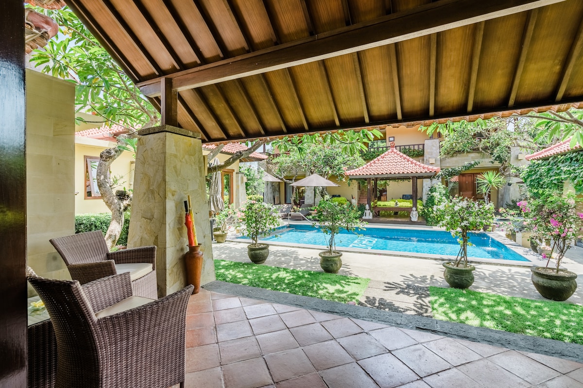 A covered outdoor seating area features two woven chairs overlooking a lush garden and a clear blue swimming pool. Surrounding greenery adds a serene atmosphere, while a sun umbrella provides shaded seating by the pool. The patio is highlighted by terracotta tiles.