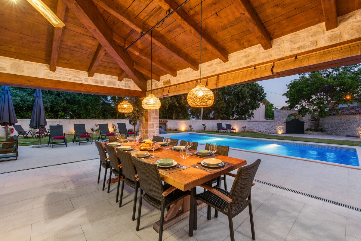 An outdoor dining area is featured under a wooden beam ceiling, with a large table set for multiple guests. Soft lighting fixtures hang above, and views of a swimming pool and lounge chairs complete the scene. Surrounding greenery adds to the tranquil environment.