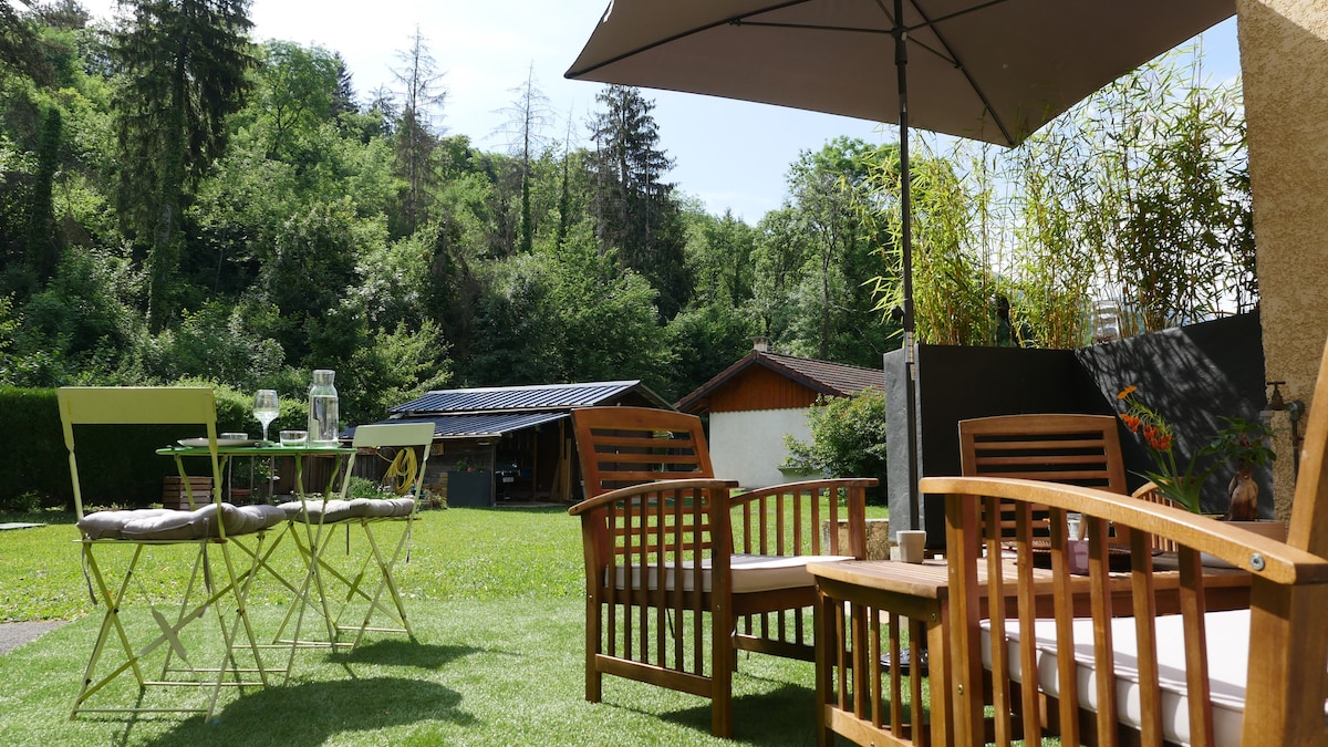 A wooden terrace area is furnished with comfortable seating and a small dining table. The space overlooks a green lawn surrounded by trees. A sun umbrella provides shade, and nearby structures are subtly integrated into the natural landscape.