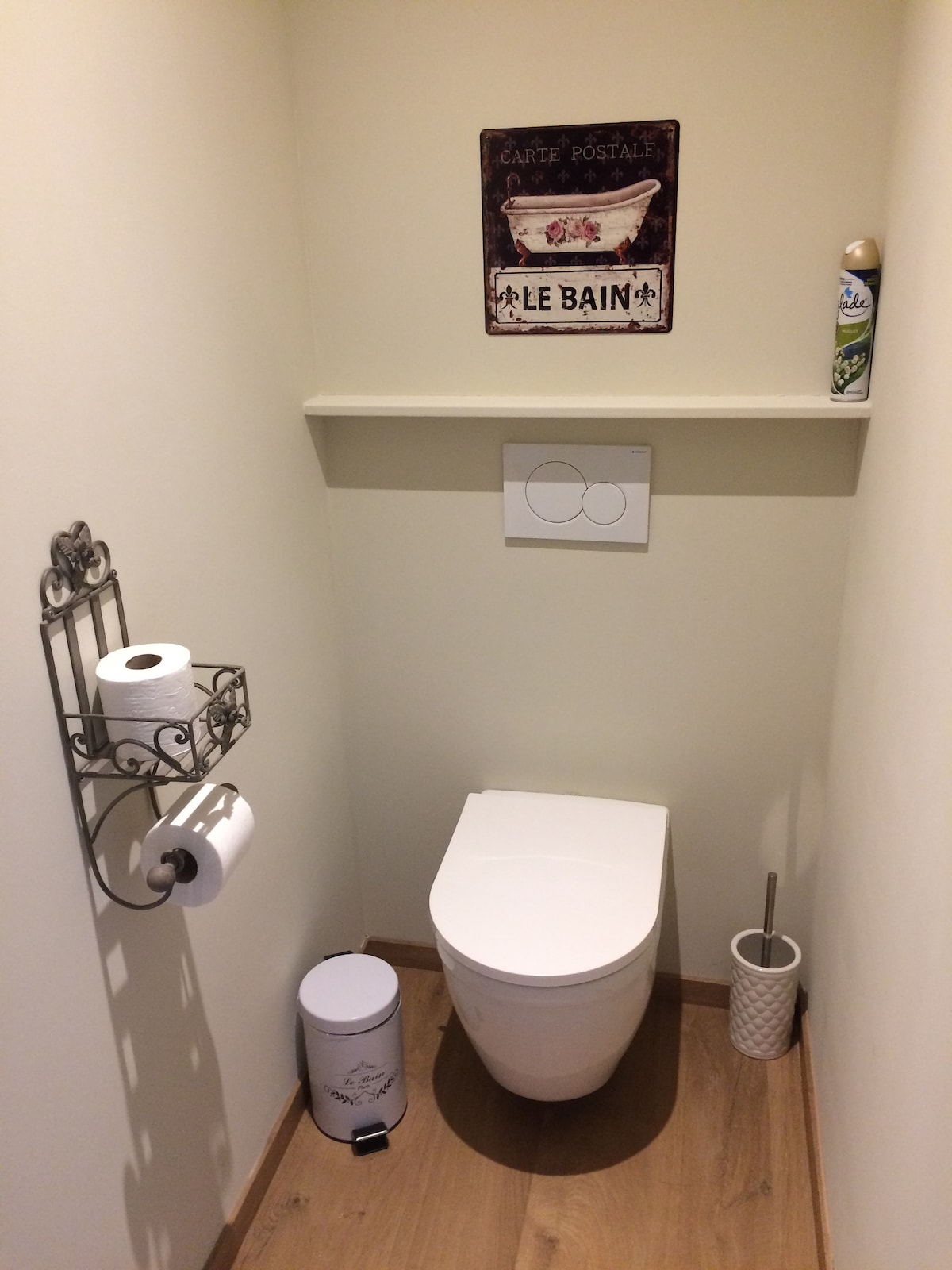 A compact bathroom space is shown, featuring a modern toilet with a dual flush system. A decorative shelf displays vintage-style signage labeled 'Le Bain.' An elegant toilet paper holder and a small waste bin are positioned nearby, enhancing the functionality of the area.