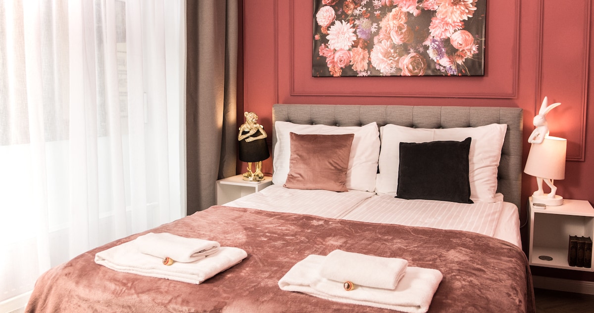A spacious bed is centered against a warm-colored wall, adorned with floral artwork. Soft towels are neatly placed at the foot of the bed, and bedside tables with decorative lamps provide ambient light. Natural light filters through sheer curtains, enhancing the inviting atmosphere.