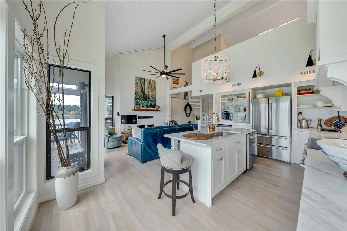 The living area features modern furnishings with a blue leather sofa, a wooden dining table, and a fully equipped kitchen. Large windows allow natural light to fill the space, showcasing views of the lake. A decorative vase with branches adds a touch of elegance.