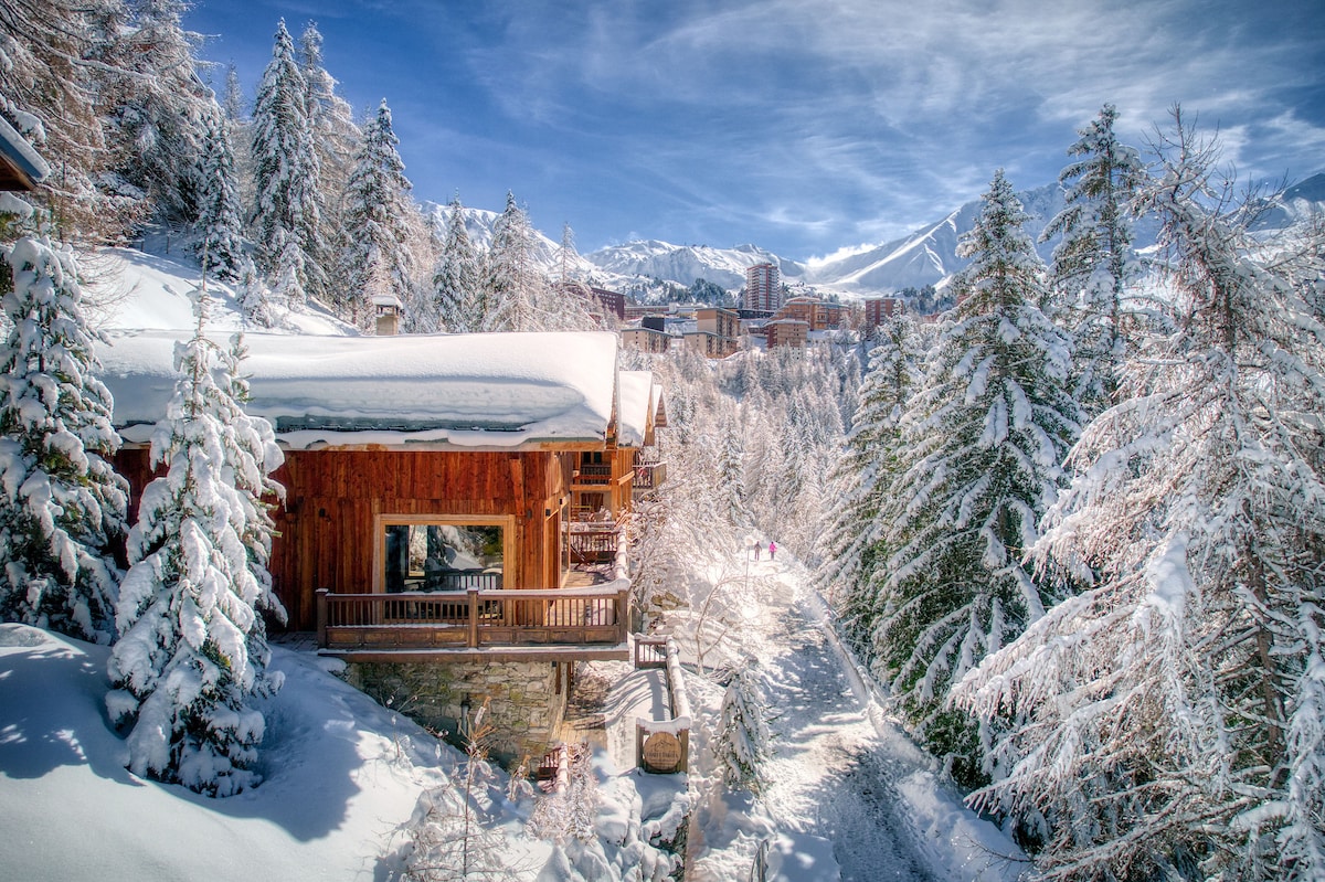 A large chalet is surrounded by snowy trees, showcasing its wooden exterior and a spacious balcony. A clear blue sky contrasts with the white snow, while ski trails can be seen winding through the picturesque landscape. The setting highlights the chalet's serene winter location.