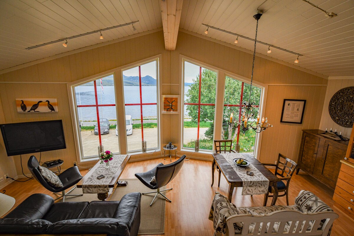 The living area is characterized by large, triangular windows that provide views of the fjord and surrounding landscape. A combination of seating options includes modern chairs and a classic wooden sofa. A dining table is set for two, complemented by decorative elements and natural light.