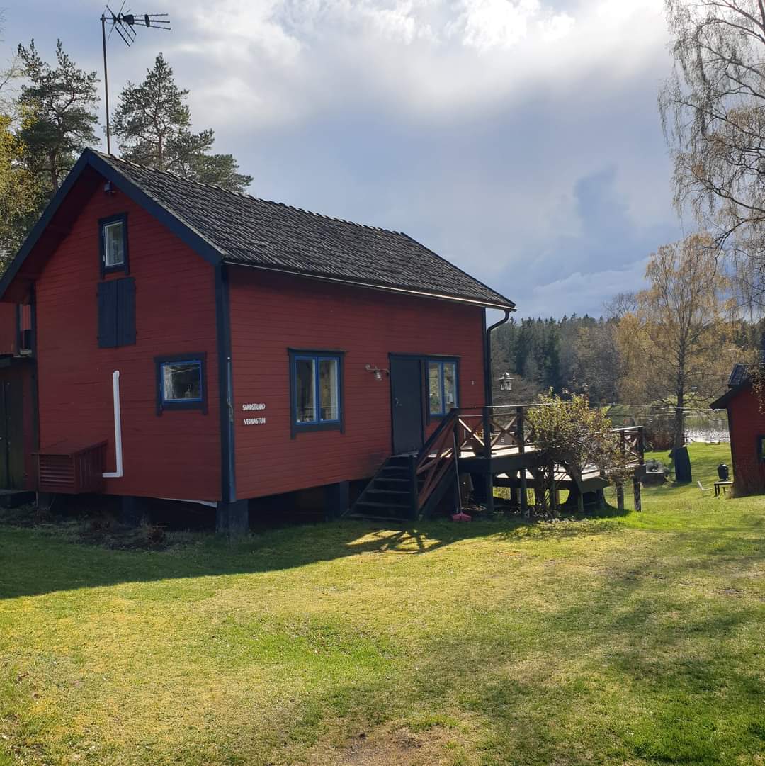 A red two-story house is nestled among trees, with a sloping lawn leading to the entrance. Stairs provide access to a wooden deck that extends from the house, inviting outdoor relaxation. The backdrop features a tranquil landscape with hints of water visible in the distance.
