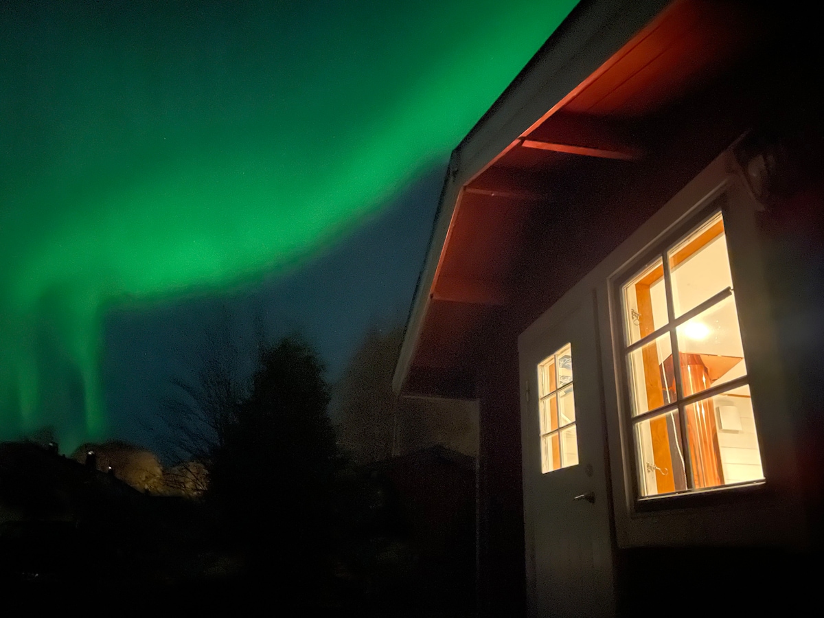 The exterior of the cabin is illuminated by artificial light, with a warm glow visible through the windows. The vibrant green hues of the northern lights dance above, creating a striking contrast against the dark sky.
