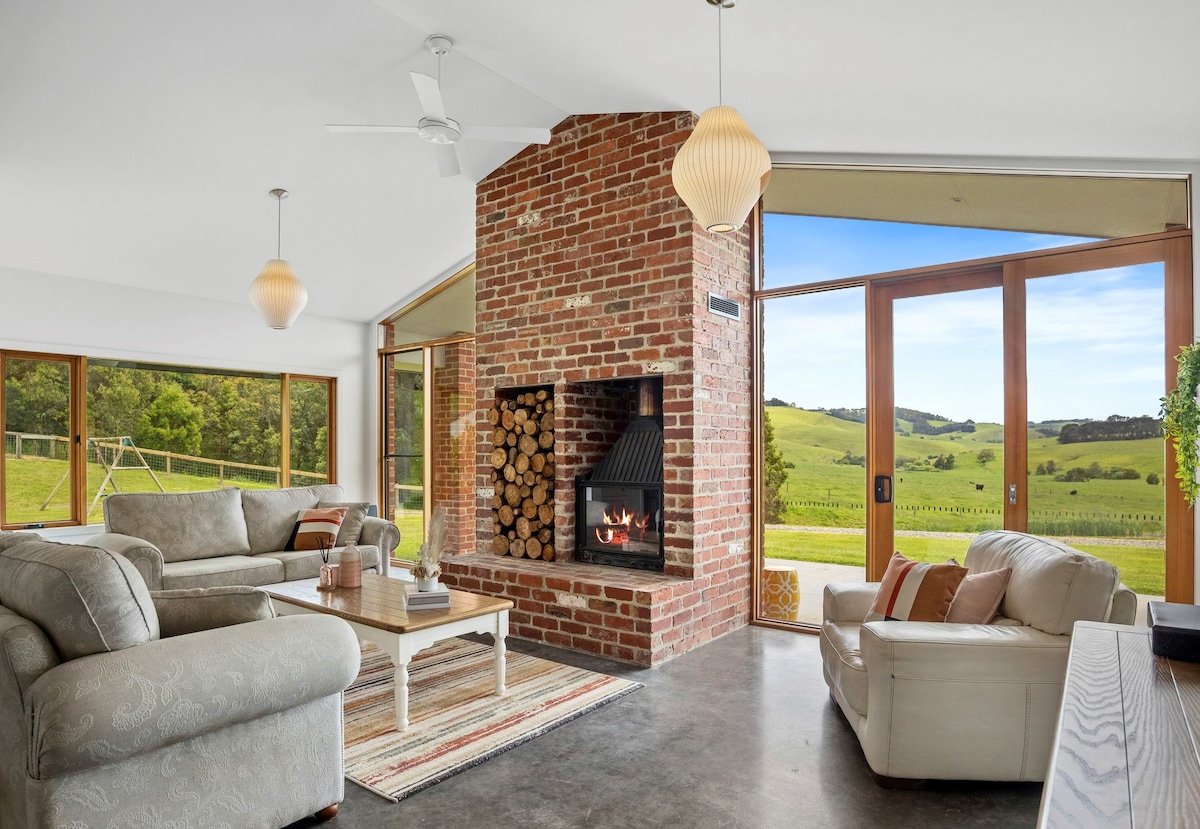 A modern living area features a brick fireplace with a wood storage area, framed by large glass doors that open to scenic views of rolling hills. Comfortable seating arrangements are complemented by natural light streaming in from multiple angles.