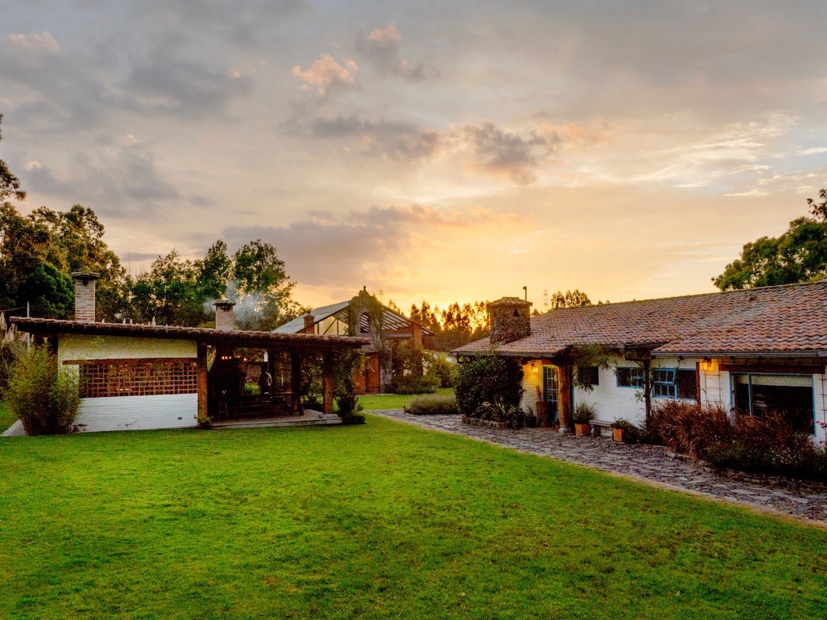 A spacious outdoor area features a well-maintained lawn, bordered by pathways and trees. Two distinct buildings are visible, one with a rustic design and the other with a welcoming porch. A sunset sky creates a serene backdrop, highlighting the tranquil countryside setting.