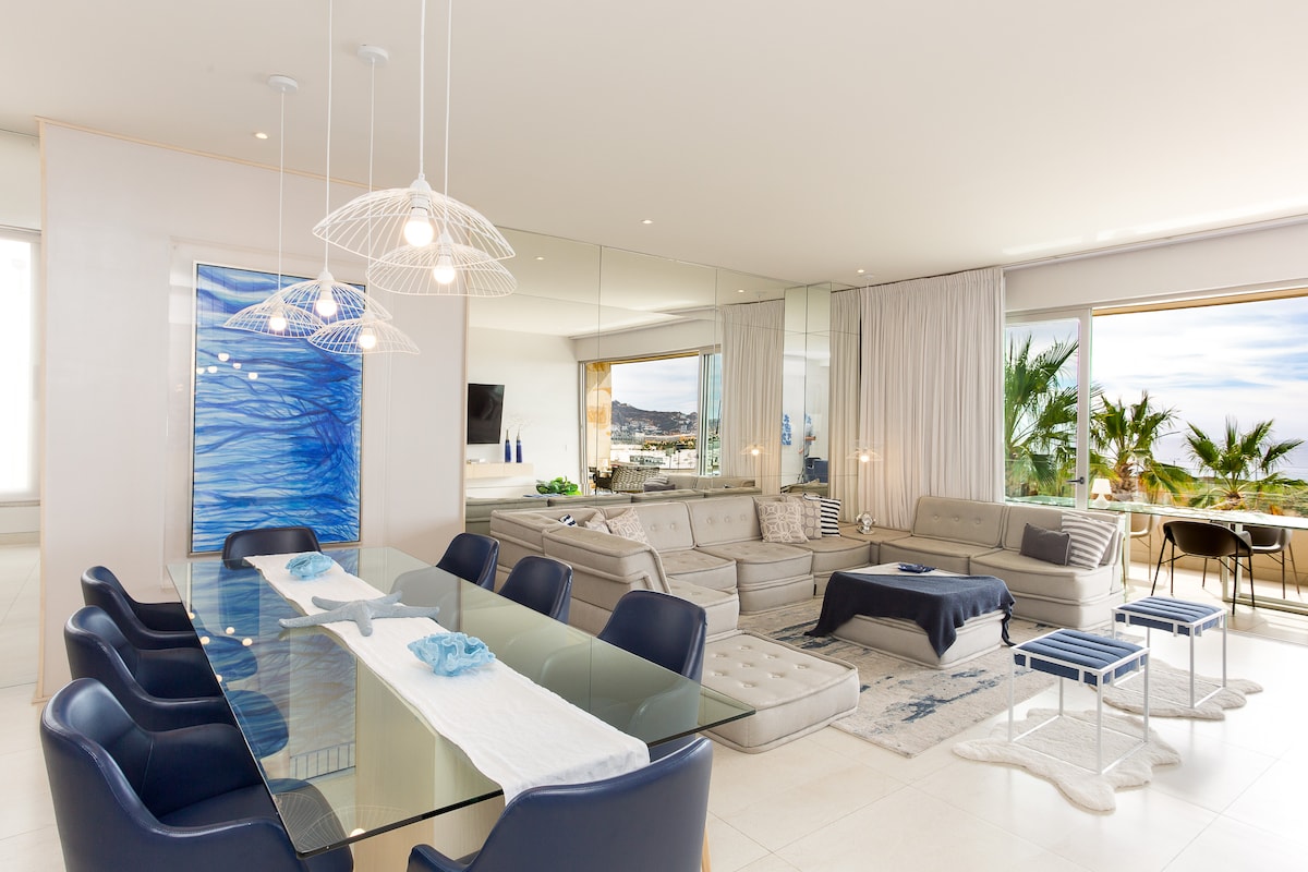 An open-concept living and dining area is shown, featuring a glass dining table surrounded by blue chairs. A comfortable seating arrangement, including light sofas and a decorative rug, is highlighted. Large windows allow for abundant natural light and offer views of greenery outside.