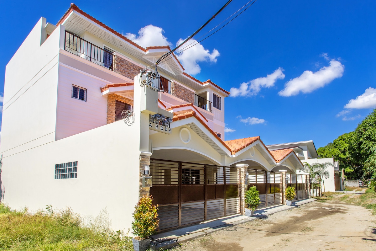 The exterior of a newly constructed villa is shown, featuring a clean design with a mix of white and stone accents. A spacious driveway is visible, flanked by low shrubs. The building is nestled under a clear blue sky, with a few clouds scattered above.