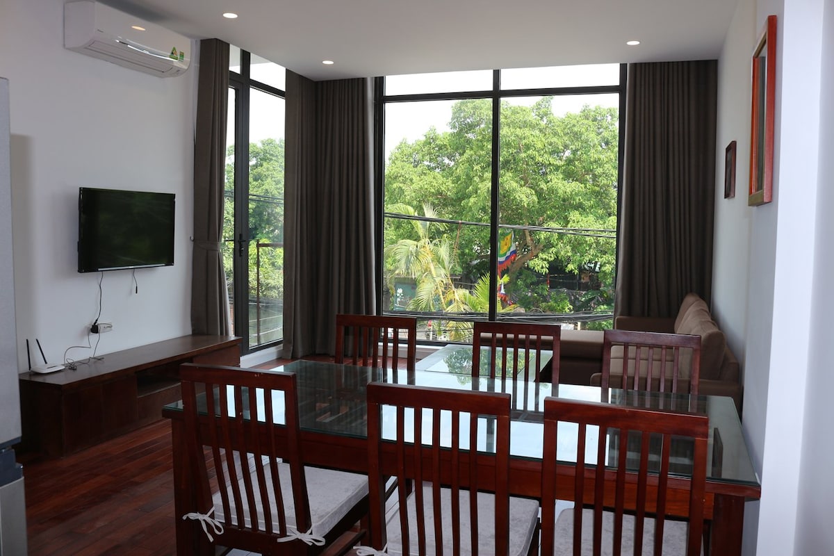 A bright and airy living space features large windows that bring in natural light and offer views of surrounding greenery. A wooden dining table is complemented by several chairs, while a cozy seating area with a sofa invites relaxation.